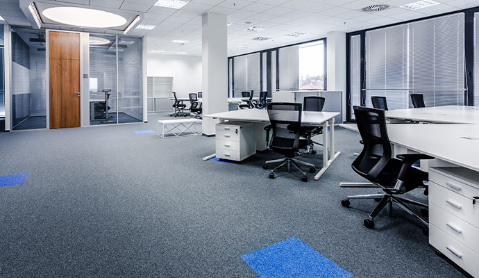 An office space featuring clean grey carpeting with blue accents, white desks, black chairs