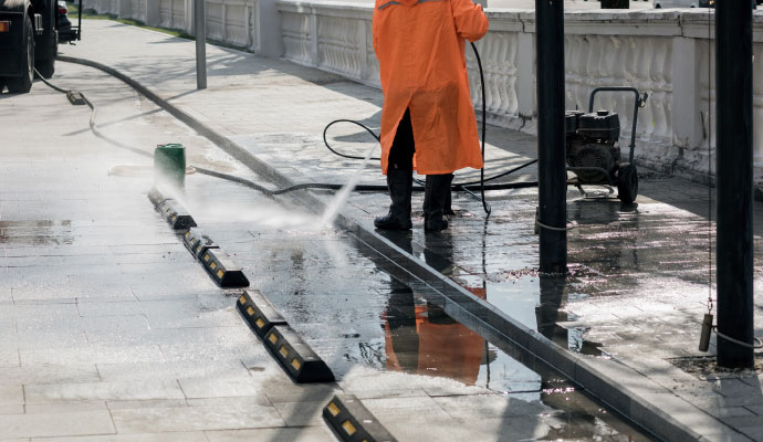 Person using high-pressure water sprayer to clean a walkway next to a parking area